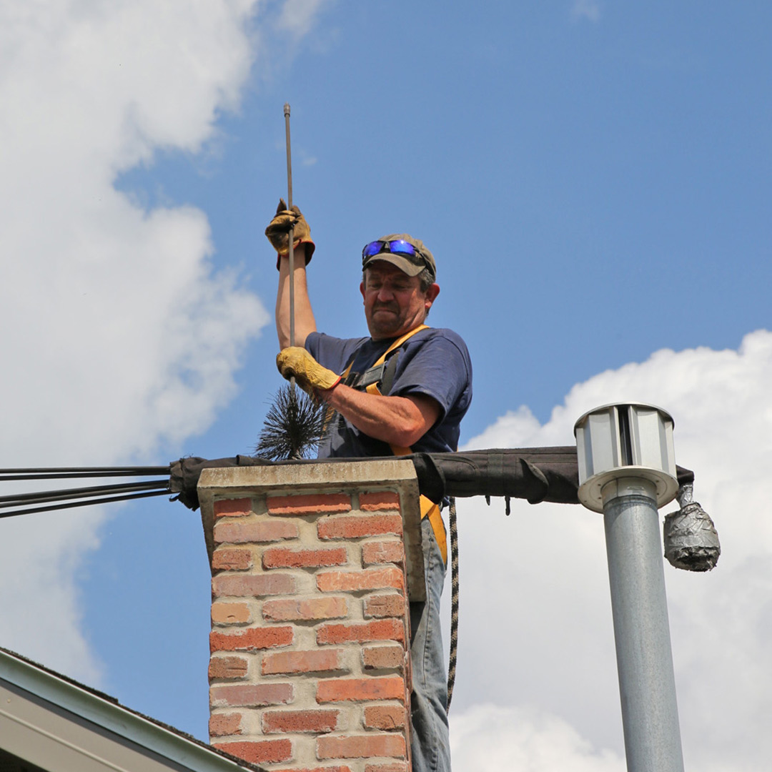 Chimney sweep in safety gear using a round brush and rod to clean a brick chimney above a roof