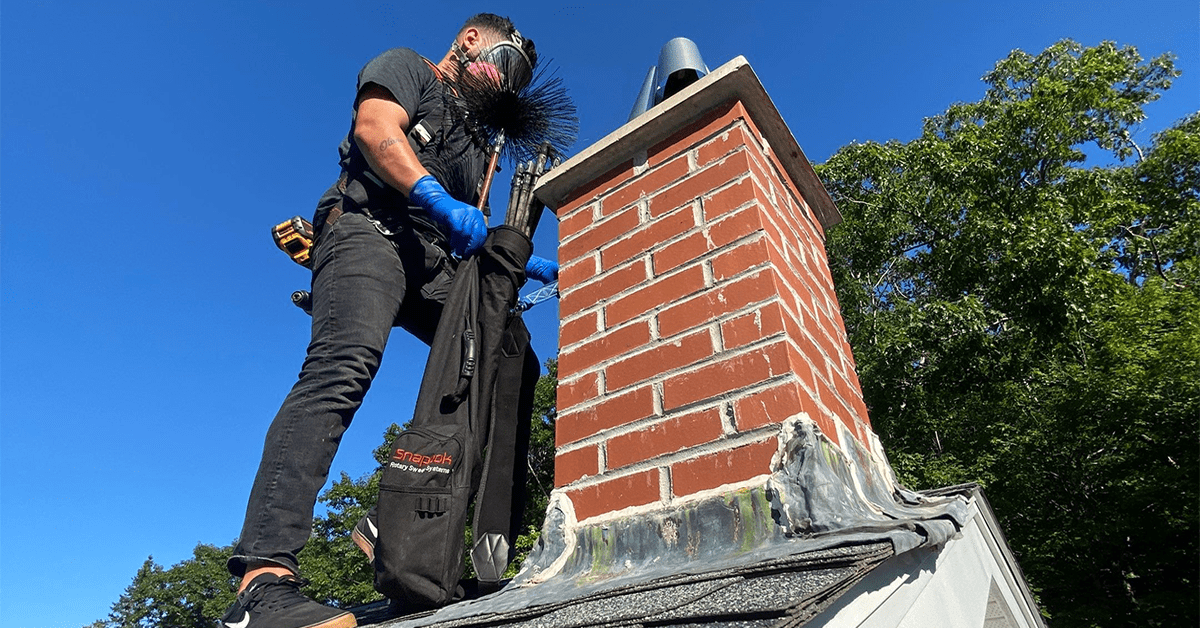 Masked chimney-sweeping technician brushing a red-brick chimney on a rooftop under a blue sky