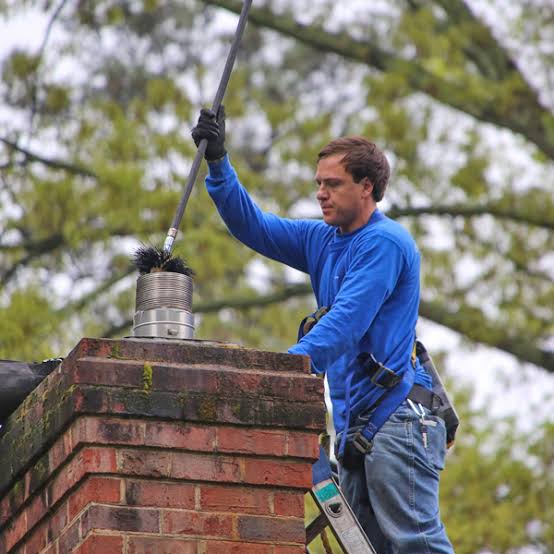 Chimney technician in a blue shirt and safety harness sweeping a brick chimney with a long-handled brush