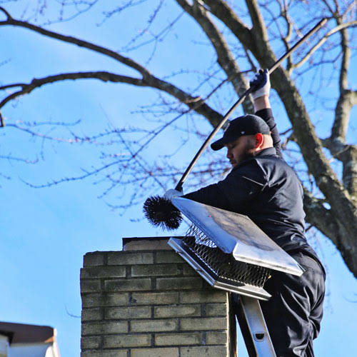 Person on a ladder brushing a brick chimney beneath a metal chimney cap on a sunny day
